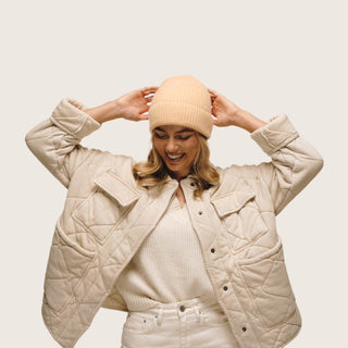 Woman adjusting Will & Bear Bambi Apricot beanie playfully, showcasing soft stretch and comfort of merino wool.