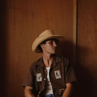 Man wearing a straw hat and brown shirt with patches against a wooden background