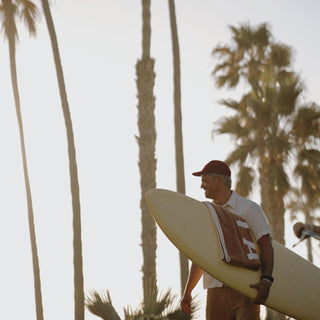Man wearing Will & Bear Archie Maroon 5-Panel Vintage Cap on a coastal adventure with surfboard and palm trees