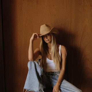 Woman wearing a straw hat and white tank top against a warm brown wall.