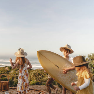Children in Will & Bear Australian Wool Kids Fedora – Little Calloway Cream walking with a surfboard near the ocean.