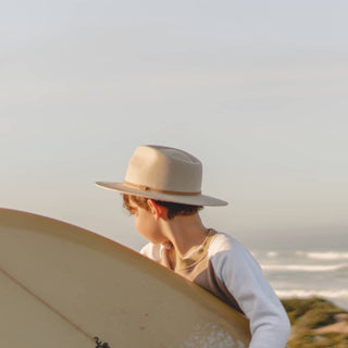 Boy wearing Will & Bear Little Calloway Cream Fedora carrying a surfboard, walking along the coast.