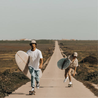 Two Men Wearing Will & Bear Beige Cord Dad Caps – Skateboarding Down a Country Road with Surfboards in Hand.