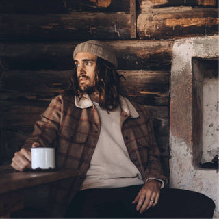 Man sitting inside a rustic cabin wearing the Will & Bear Doc Fawn Wool Fisherman’s Beanie, holding a mug near the fireplace.