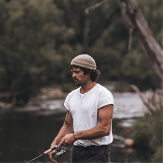 Man fishing by a calm river, wearing the Will & Bear Doc Fawn Wool Fisherman’s Beanie and white t-shirt.