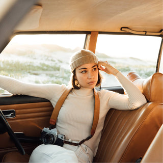 Woman seated inside a classic car window seat, wearing the Will & Bear Doc Fawn Wool Fisherman’s Beanie with camera in lap.