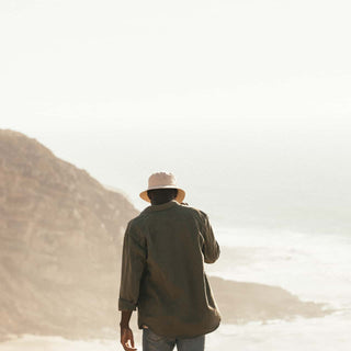 Man walking toward the ocean in the Will & Bear Fischer Bone White Bucket Hat, rugged cliffs in the distance.