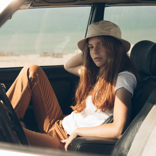 Woman relaxing in car seat wearing the Will & Bear Fischer Bone White Bucket Hat during a coastal road trip.