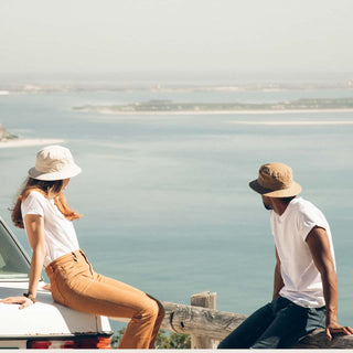 Man and woman wearing Will & Bear Fischer Sand Waxed Canvas Bucket Hats, sitting on a sea wall overlooking the ocean.