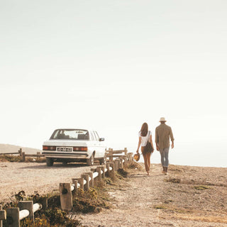 Couple walking on a coastal path with a vintage car in the background, both wearing Will & Bear bucket hats including Fischer Sand.