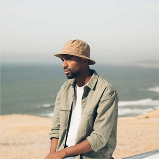 Man wearing the Will & Bear Fischer Sand Waxed Canvas Bucket Hat standing near the ocean on a coastal cliff.