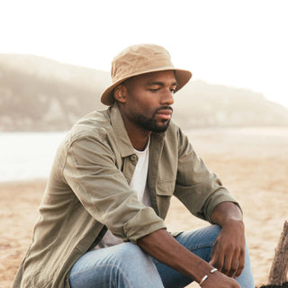 Man seated by the water in Will & Bear Fischer Sand Waxed Canvas Bucket Hat, wearing green overshirt and jeans.