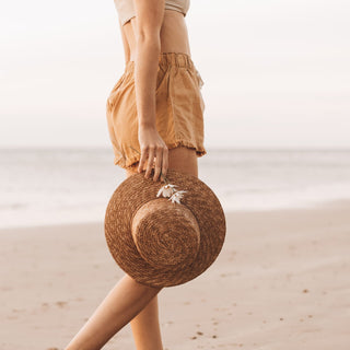 Woman walking along the beach holding the Will & Bear Harvey Amber Women’s Straw Boater by the brim.