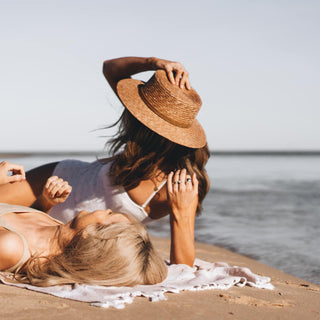 Two women lounging by the beach wearing the Will & Bear Harvey Amber Women’s Straw Boater under the afternoon sun.