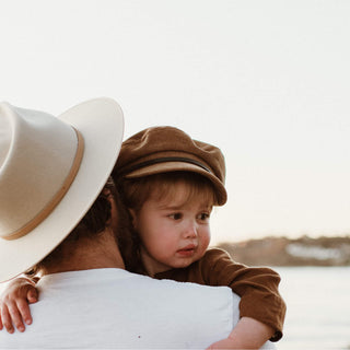 Small child wearing Will & Bear Kids Newsboy Cap – Little Baker Tan hugging adult on a sandy beach.