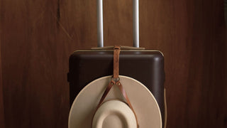 Leather hat holder securing a wide brim hat to a suitcase handle