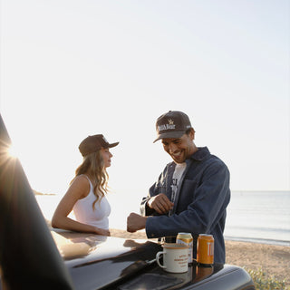 Man in a Tanner Brown 5 panel cap laughing by a car bonnet on the beach, woman in a Carter Brown trucker cap beside him