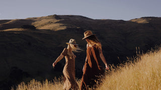 Two friends wander through a sunlit field, one in a dress and a floppy hat, immersed in nature.