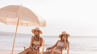 Two women relaxing under an umbrella, wearing beach hats by Will & Bear, the ocean behind them.