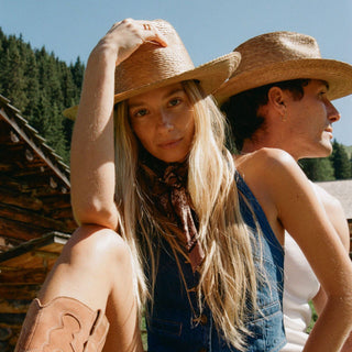 Two women outdoors in front of a rustic wooden building and trees, wearing straw cowboy hats, denim overalls, and cowboy boots. One woman faces forward while adjusting her hat, and the other is turned to the side in profile.