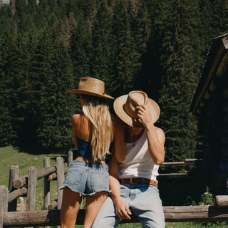 A couple wearing straw cowboy hats sits on a rustic wooden fence in a sunny mountain setting with a forest background.