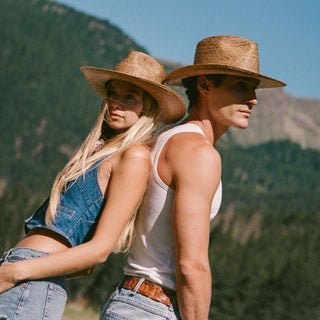 A man and a woman wearing straw western  hats pose back-to-back outdoors against a mountain backdrop. The woman wears a denim vest and jeans, and the man wears a white tank top and jeans.