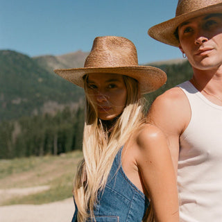 A lifestyle shot of a couple outdoors wearing matching natural straw wide-brimmed hats. The woman in the foreground wears a straw hat and denim overalls, while the man behind her wears a white tank top and a matching straw hat, set against a scenic mountain background.