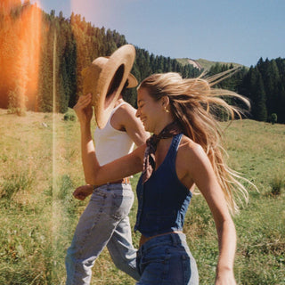 Two women running and laughing in a grassy outdoor field with trees and mountains in the background. One wears a straw hat, a white tank top, and light blue jeans, while the other wears a dark blue sleeveless top and denim bottoms.