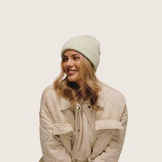 Woman sitting front on and staring away from the camera wearing Bambi Sage Green knit beanie