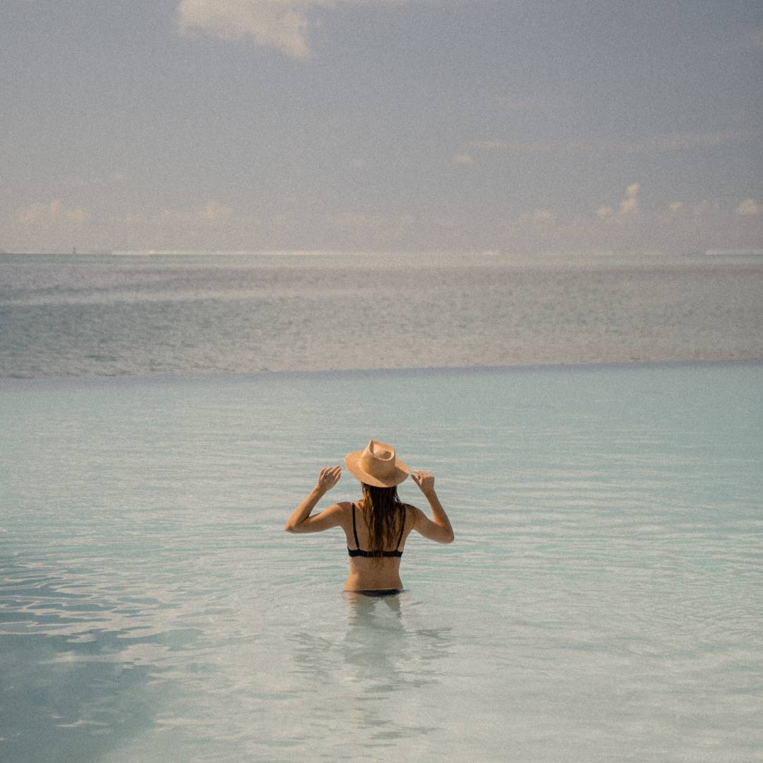 A woman with long blonde hair wearing a wide brim straw hat, she is standing in the ocean.