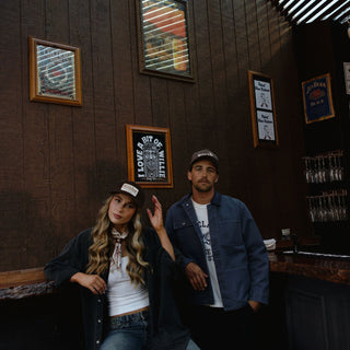 Two people leaning at a dimly lit bar counter with framed wall art behind them. One wearing Carter Brown trucker hat the other wearing Tanner Brown 5-panel cap