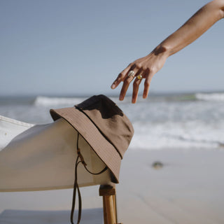 Will & Bear removable string bucket hat hanging off a surfboard by the ocean.