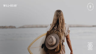 Women at the beach carrying a surfboard and hat clipped to her bag