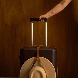 Leather hat holder securing hat to rolling luggage while traveling