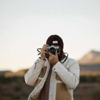 Person wearing the Levi Dark Brown beanie holding a vintage camera in a desert setting