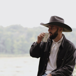 Man enjoying coffee by a lake while wearing the Will & Bear Buck Smoke black felt cowboy hat, styled for outdoor adventure.