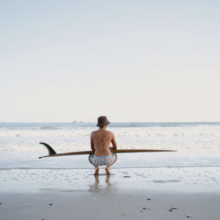 Man standing on the beach holding a surfboard wearing a string bucket hat