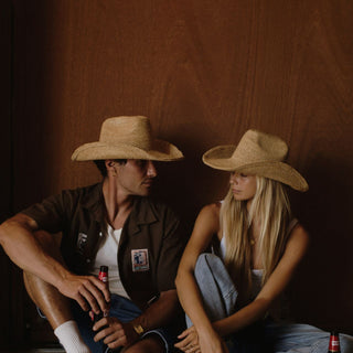 Two people wearing straw hats sitting against a wooden wall.