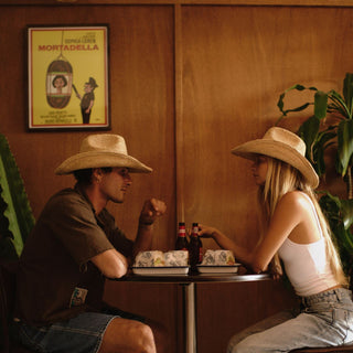 Two people in cowboy hats sitting at a table with bottles and food, in a room with a poster on the wall.