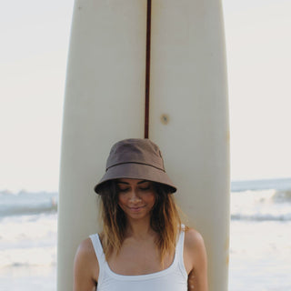 Woman wearing a brown string bucket hat standing by a surfboard