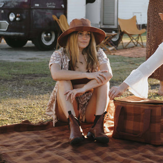 Woman sitting on a picnic blanket wearing the Will & Bear Camp Bodhi Carob recycled hemp bucket hat at a rustic campsite.