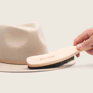 Hand using the Will & Bear Hat Brush to clean a wool felt hat, demonstrating gentle maintenance on a light-coloured brim.
