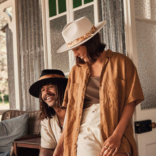 Couple on a veranda, each wearing a hat styled with the Will & Bear Hat Scarf Mustard tied around the crown.
