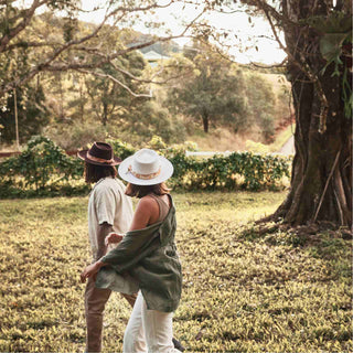 Couple walking through a sunlit field, the woman’s hat styled with the Will & Bear Hat Scarf Mustard tied around the crown.