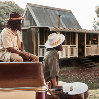 Outdoor scene with two people beside a cabin, each wearing hats styled with the Will & Bear Hat Scarf Mustard.
