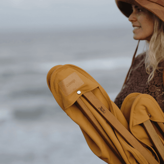 Woman carrying a folded Will & Bear x Wandering Folk Slow Road Camp Chair near the ocean, ready for a beach adventure.
