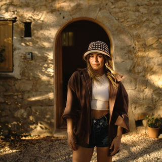 A woman standing in front of a stone building wearing a patterned crochet hat, a brown jacket, and denim shorts.