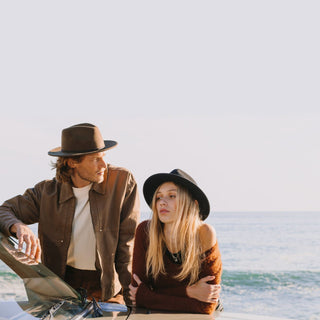 Woman wearing wide-brim fedora hat leaning on a car by the beach with a guy wearing William Brown mens wide-brim fedora hat