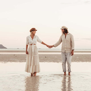 Couple holding hands wearing Will & Bear hats, including the Baker Fawn white cord newsboy cap, during a coastal walk.