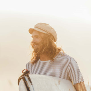 Man wearing the Will & Bear Baker Fawn white cord newsboy cap on a beach holding a surfboard.